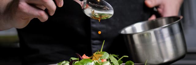 Close-up of the hands of a male chef on a black background. Pour sauce from the spoon on the salad dish. Food decoration.
